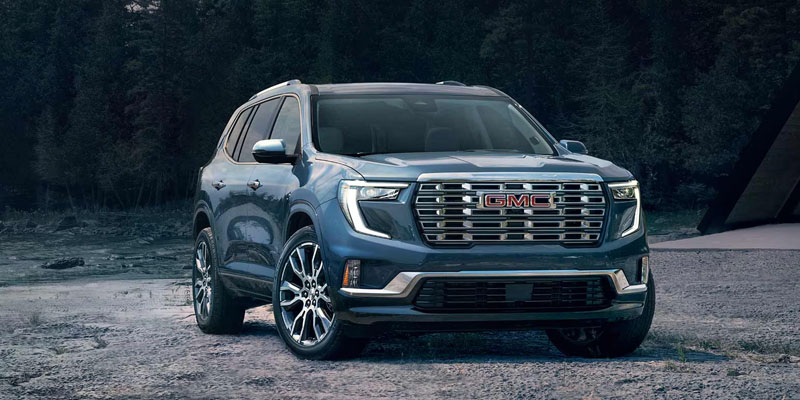 A blue GMC Acadia SUV parked on a rugged terrain, surrounded by trees in the background.